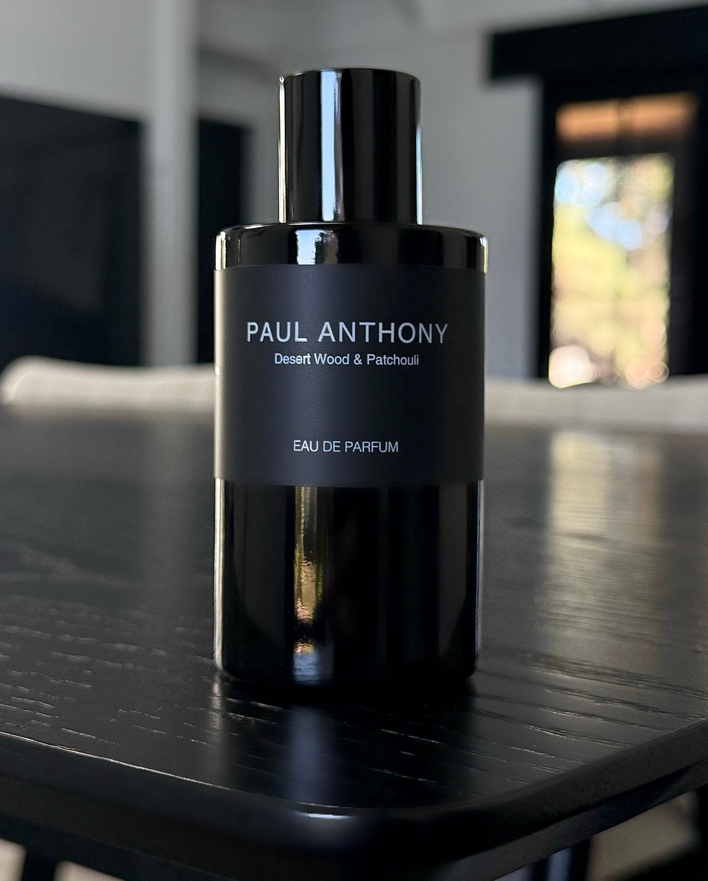 A bottle of Paul Anthony Desert Wood & Patchouli Eau de Parfum, featuring a glossy black cylindrical bottle and cap with a matte black label, set on a dark wooden table in a modern interior.
