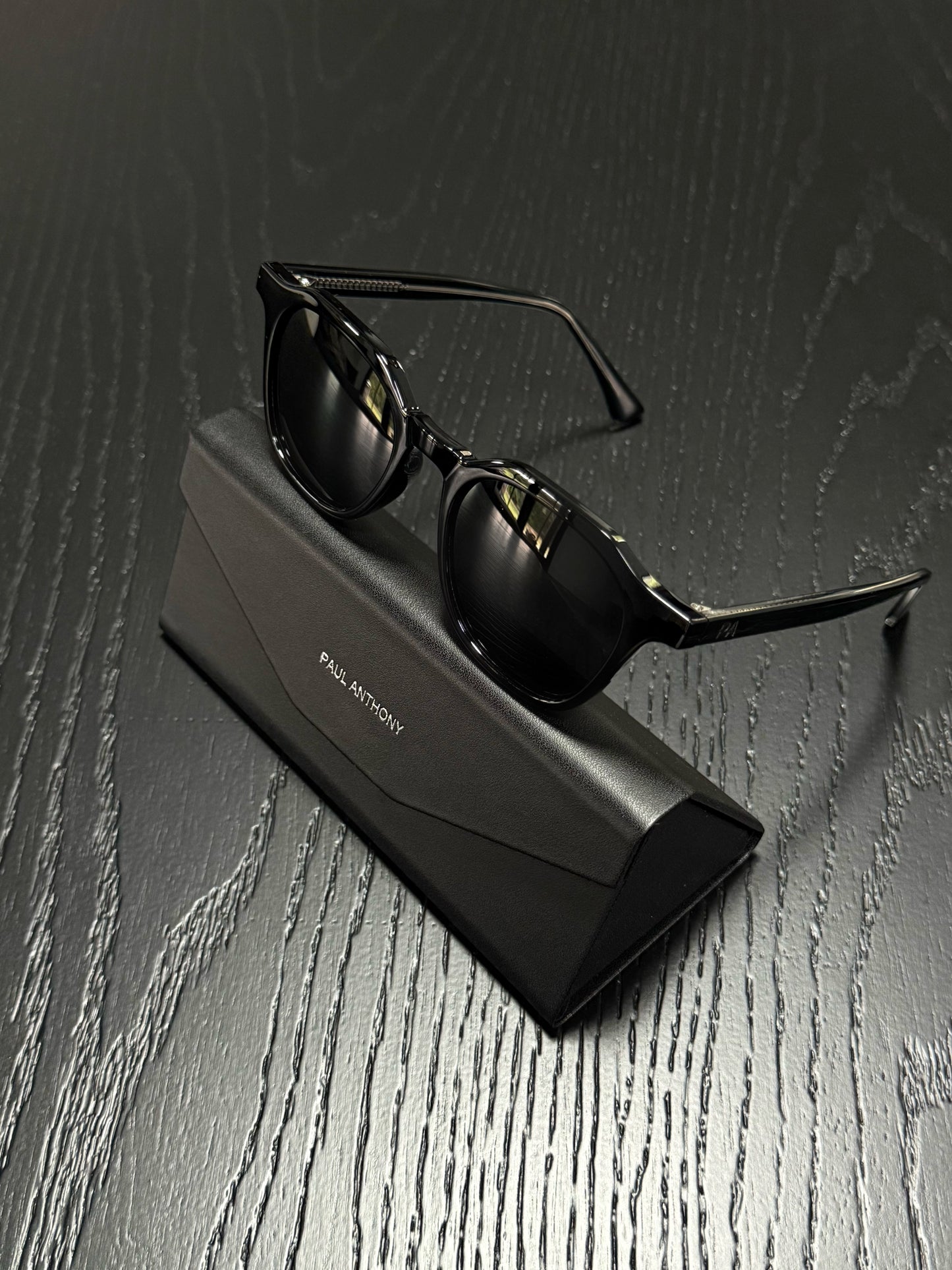 Black sunglasses with a thin metal frame resting on a black rectangular case, set on a dark wood-textured surface. The case has 'PAUL MONTROY' printed on it.