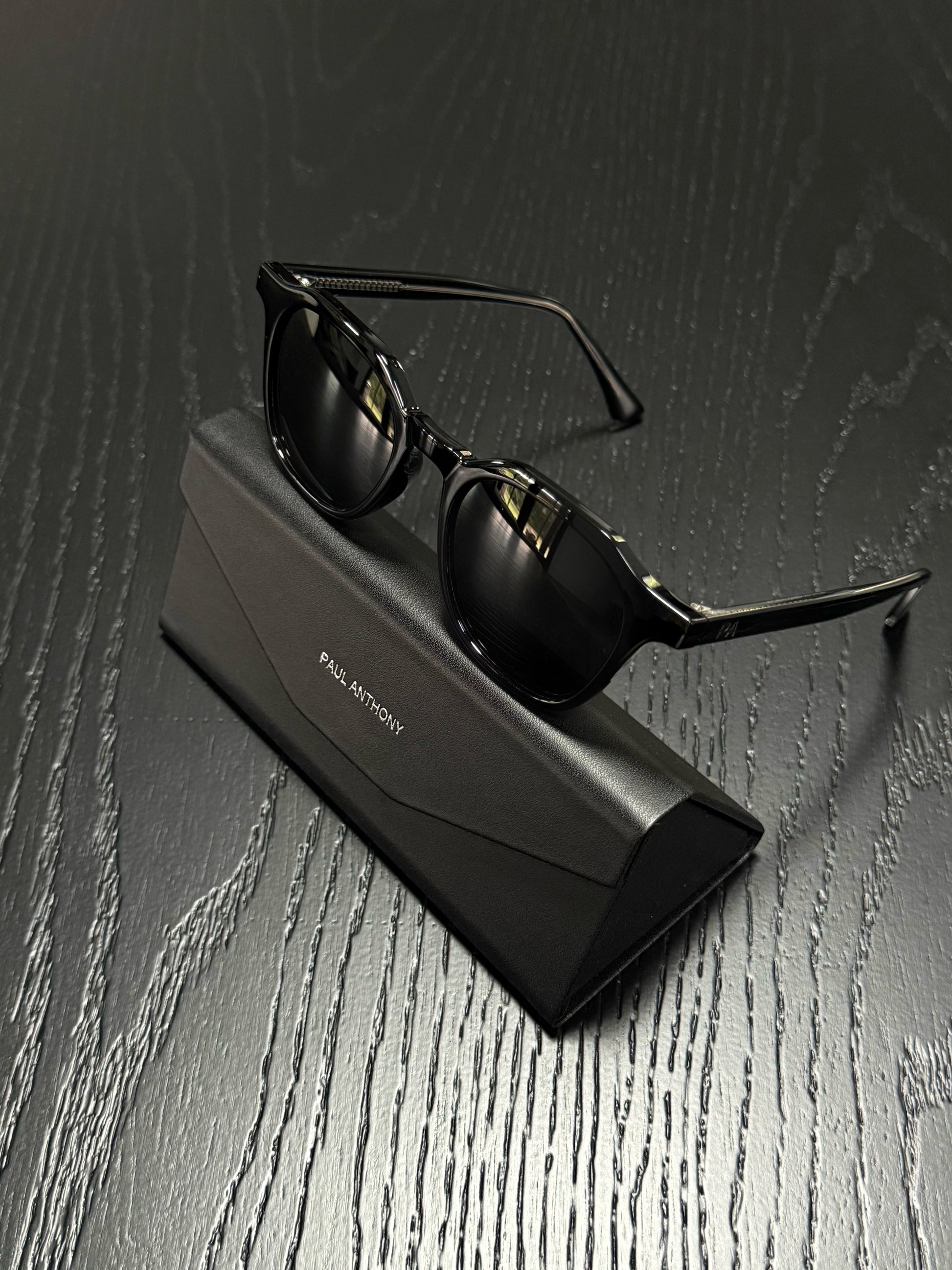 Black sunglasses with a thin metal frame resting on a black rectangular case, set on a dark wood-textured surface. The case has 'PAUL MONTROY' printed on it.
