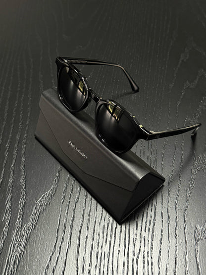 Black sunglasses with a thin metal frame resting on a black rectangular case, set on a dark wood-textured surface. The case has 'PAUL MONTROY' printed on it.