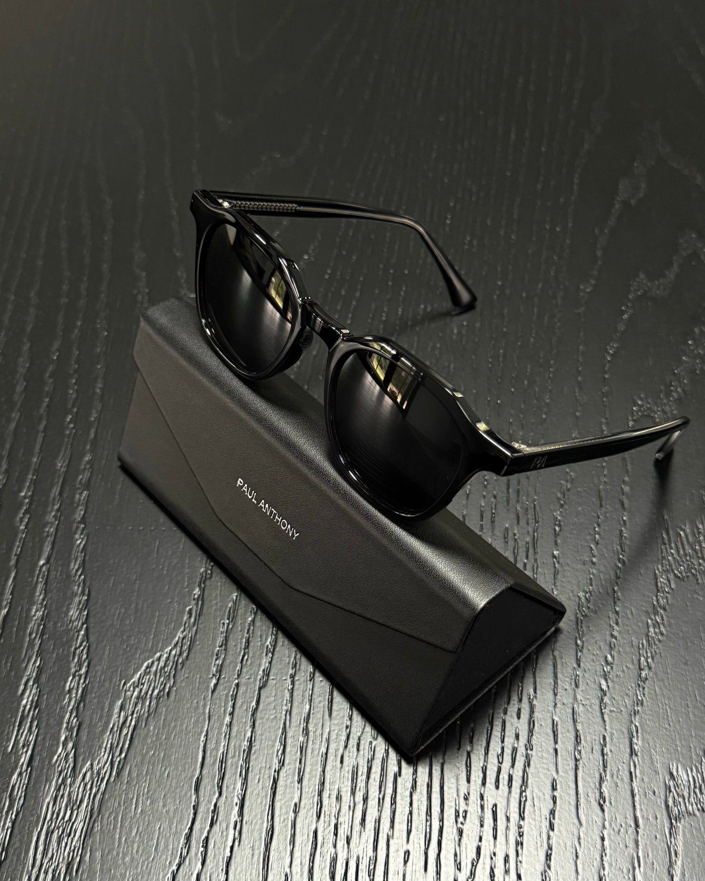 Black sunglasses with a glossy finish resting on a black Paul Martin brand case, set against a dark wood-textured background.