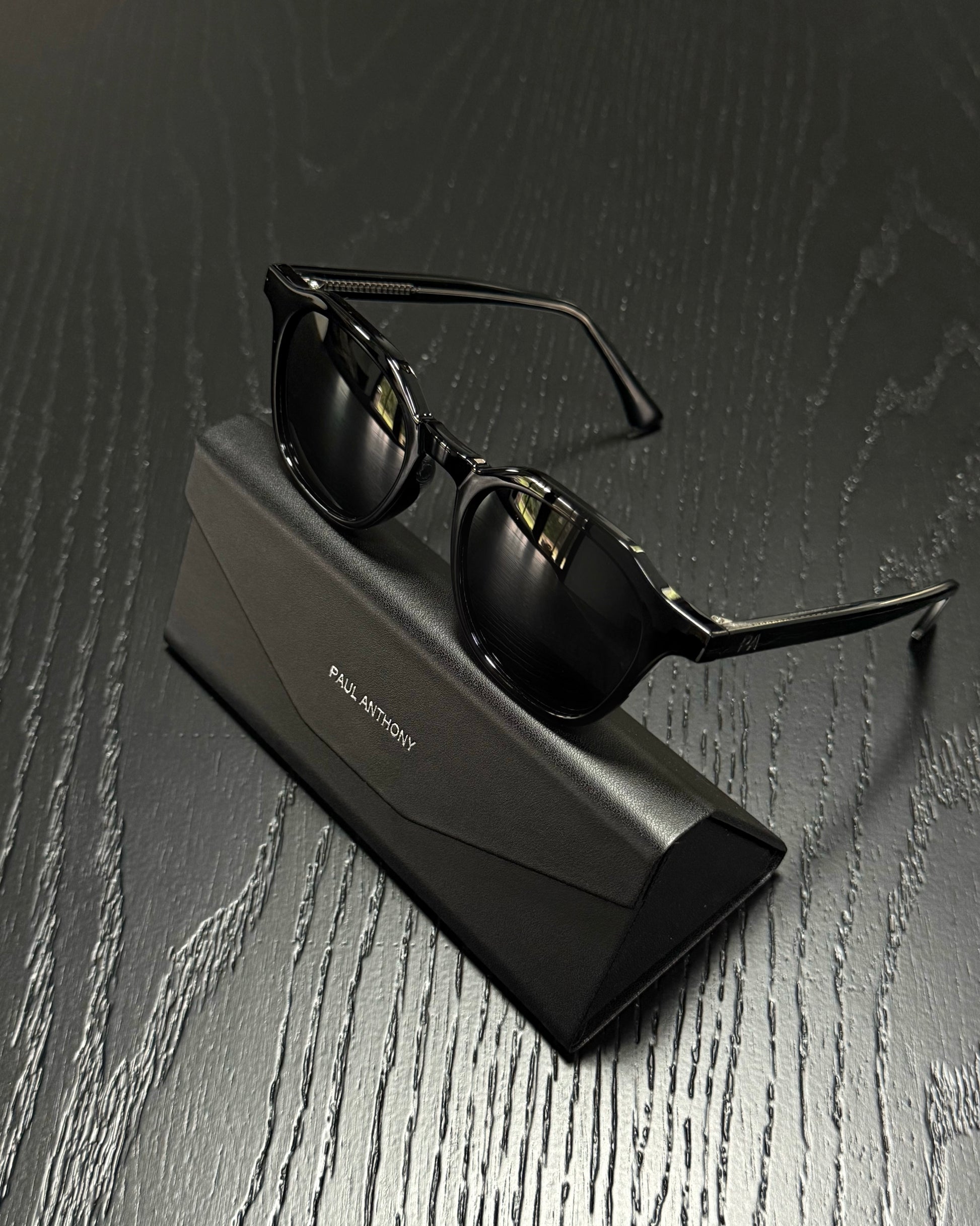 Black sunglasses with a glossy finish resting on a black Paul Martin brand case, set against a dark wood-textured background.