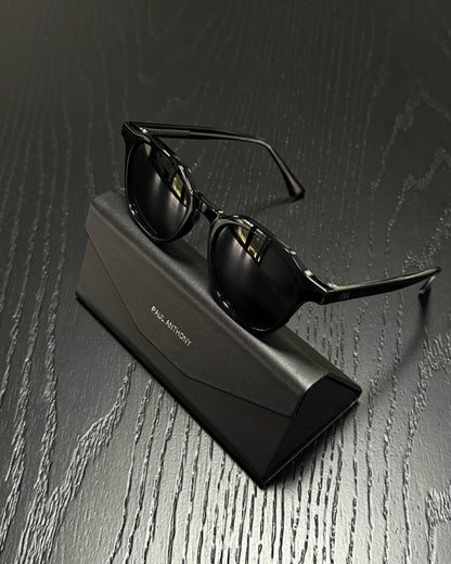 Black sunglasses with a glossy finish resting on a black Paul Martin brand case, set against a dark wood-textured background.