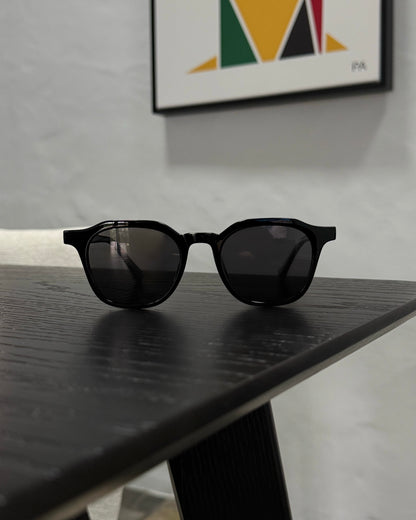 Black round-frame sunglasses with dark lenses resting on a dark wooden table, with a white wall and framed wall art in the background.