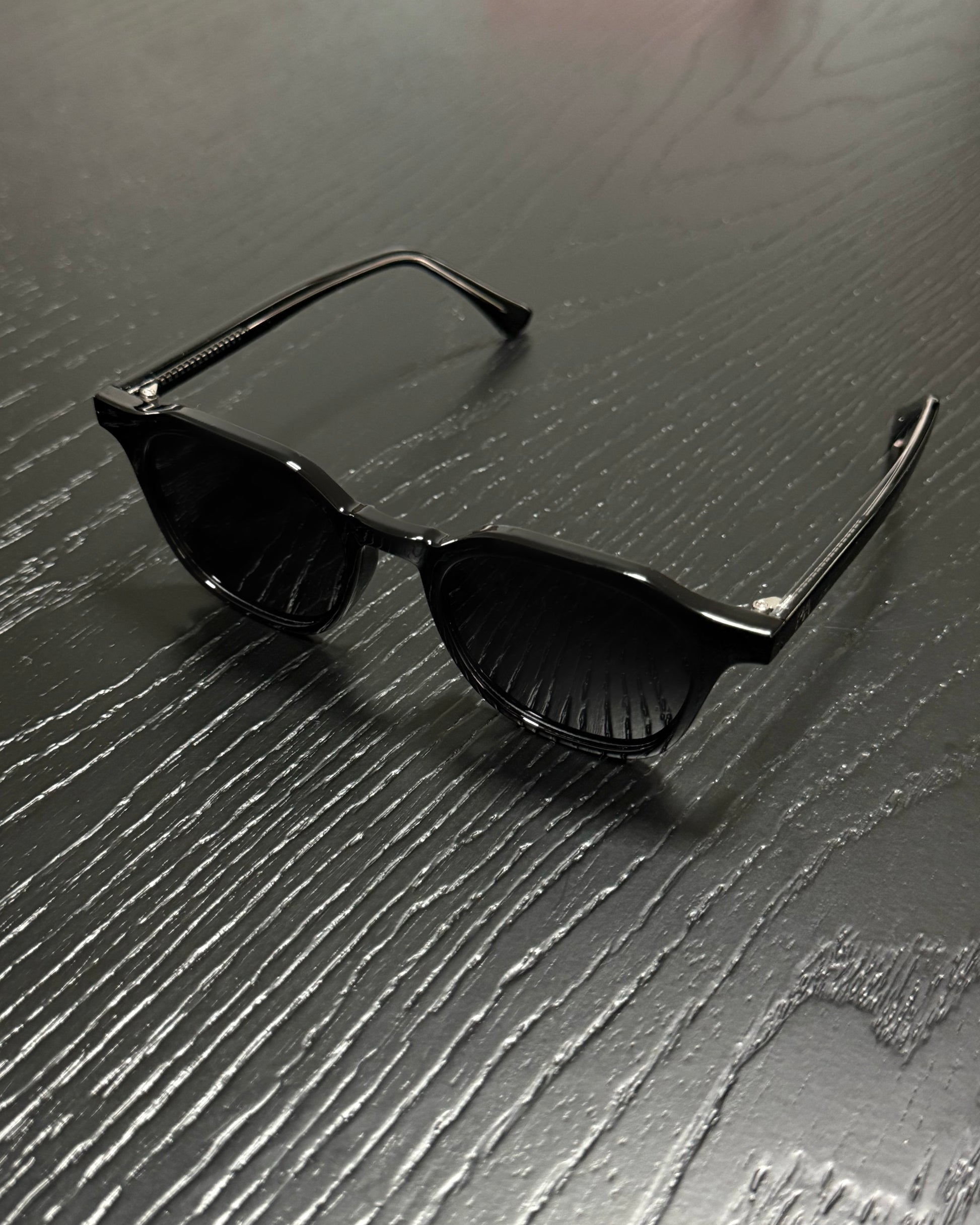 Black sunglasses with a glossy finish and dark lenses, resting on a dark gray wood-textured surface.