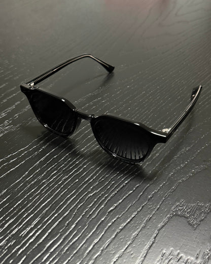 Black sunglasses with a glossy finish and dark lenses, resting on a dark gray wood-textured surface.