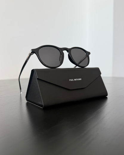 Black sunglasses on a black carrying case with 'Paul Anthony' branding, on a reflective surface.
