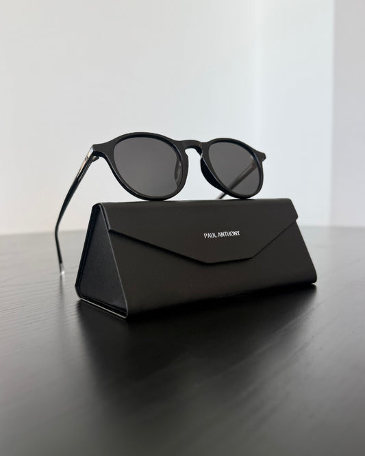 Black sunglasses on a black carrying case with 'Paul Anthony' branding, on a reflective surface.